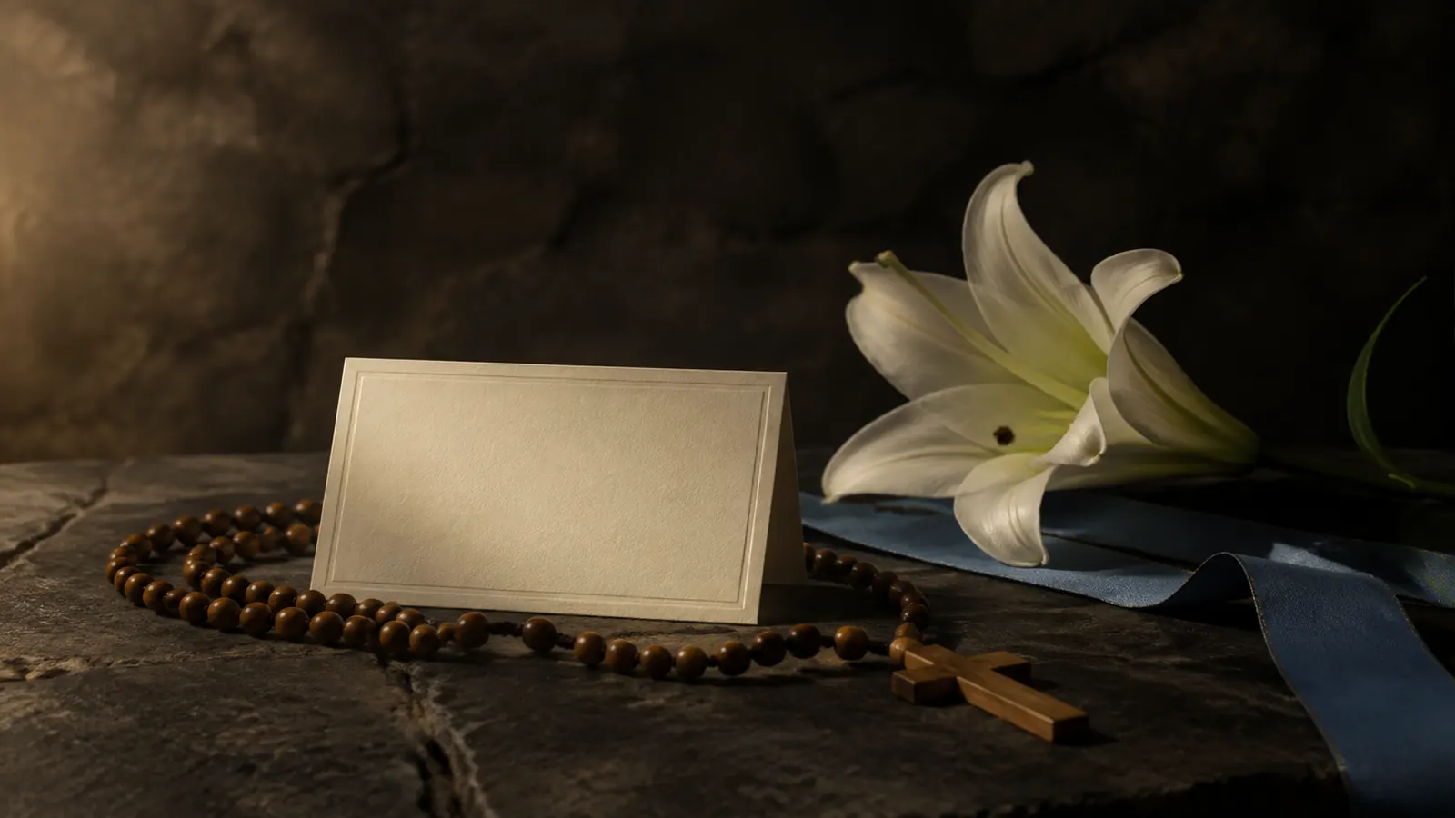 A blank ivory name card beside a wooden rosary, white lily, and muted blue ribbon on a dark stone surface